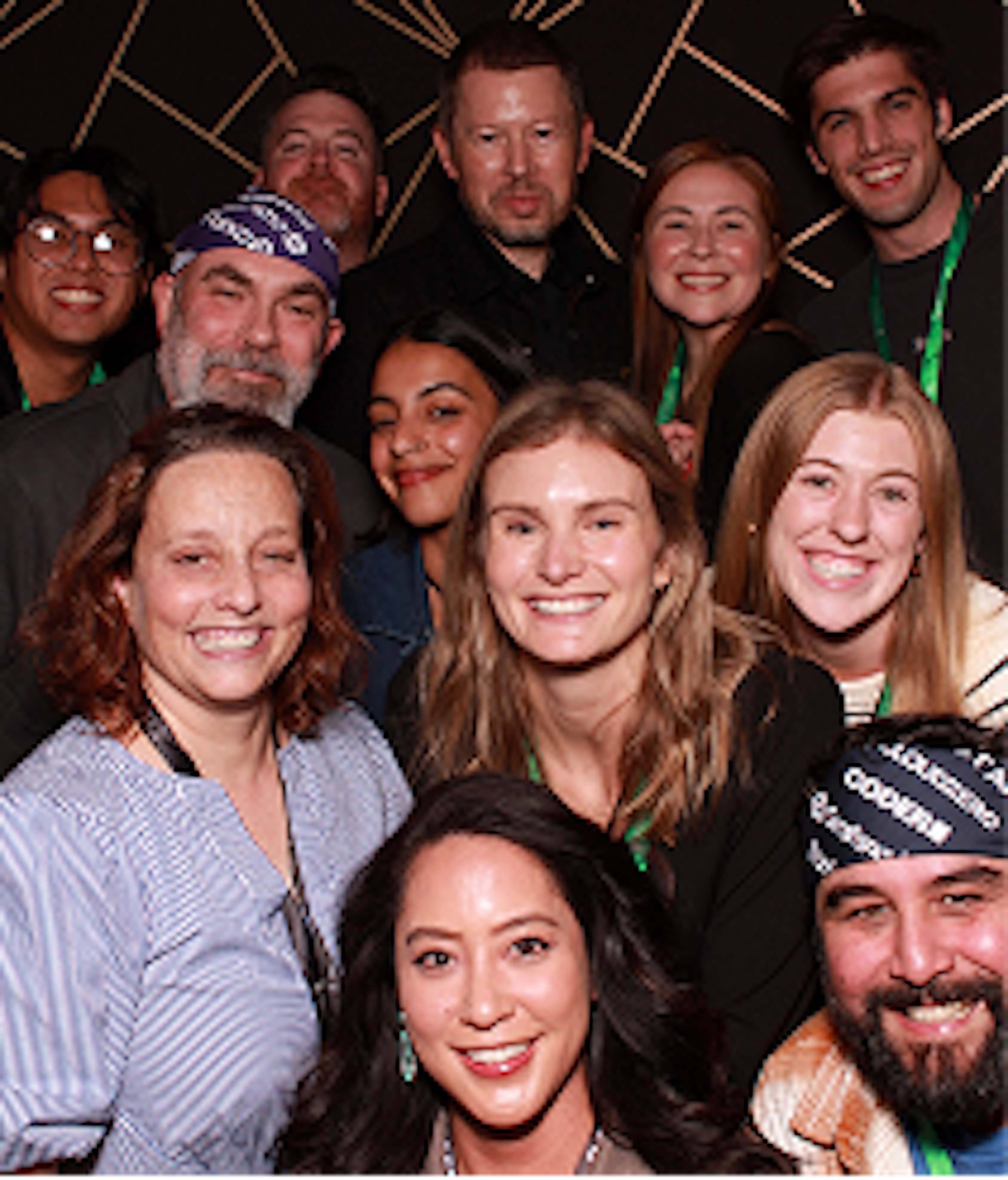 group of 12 people smiling together. 2 are wearing headbands with company logos including Coder, CloudZero, and Grafana