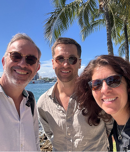 3 people wearing sunglasses, smiling beneath a palm tree and sunny blue sky