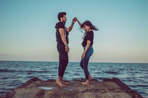 A man and woman dance barefoot at the edge of a rocky outcrop by the sea. The woman twirls under the man's arm, both smiling and enjoying the moment. They are casually dressed against a backdrop of the calm ocean and a clear blue sky.