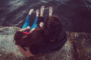 A couple is sitting on the edge of a rocky surface near a body of water. They have their feet dangling over the water, with one person resting their head on the other's shoulder. They are seen from above, creating a peaceful, intimate scene.