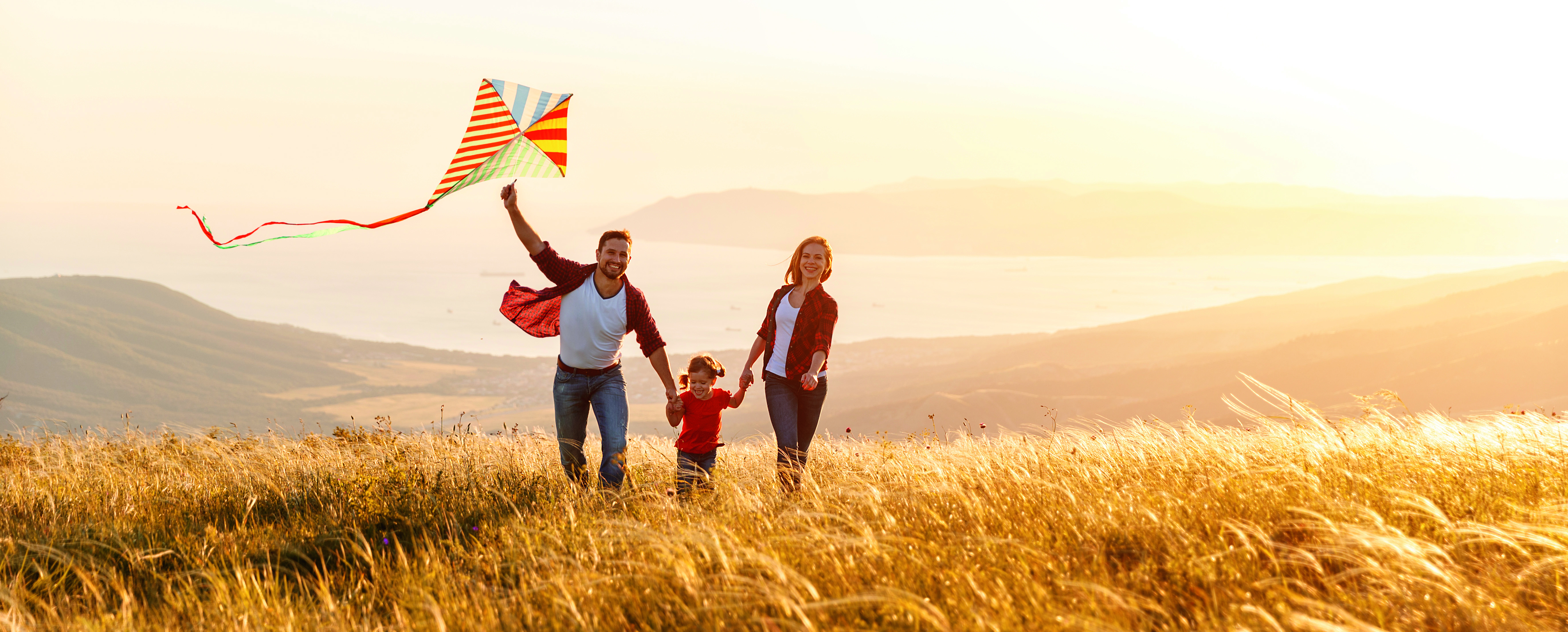 Eine glückliche Familie rennt Hand in Hand über ein golden schimmerndes Kornfeld. Der Vater hält einen Drachen in der Hand. Das Tal im Hintergrund erstrahlt in einem goldenen Licht. Mach auch Sie sich eine Freude und schenken Sie sich oder Ihren Liebsten Gold mit Golden Gates.