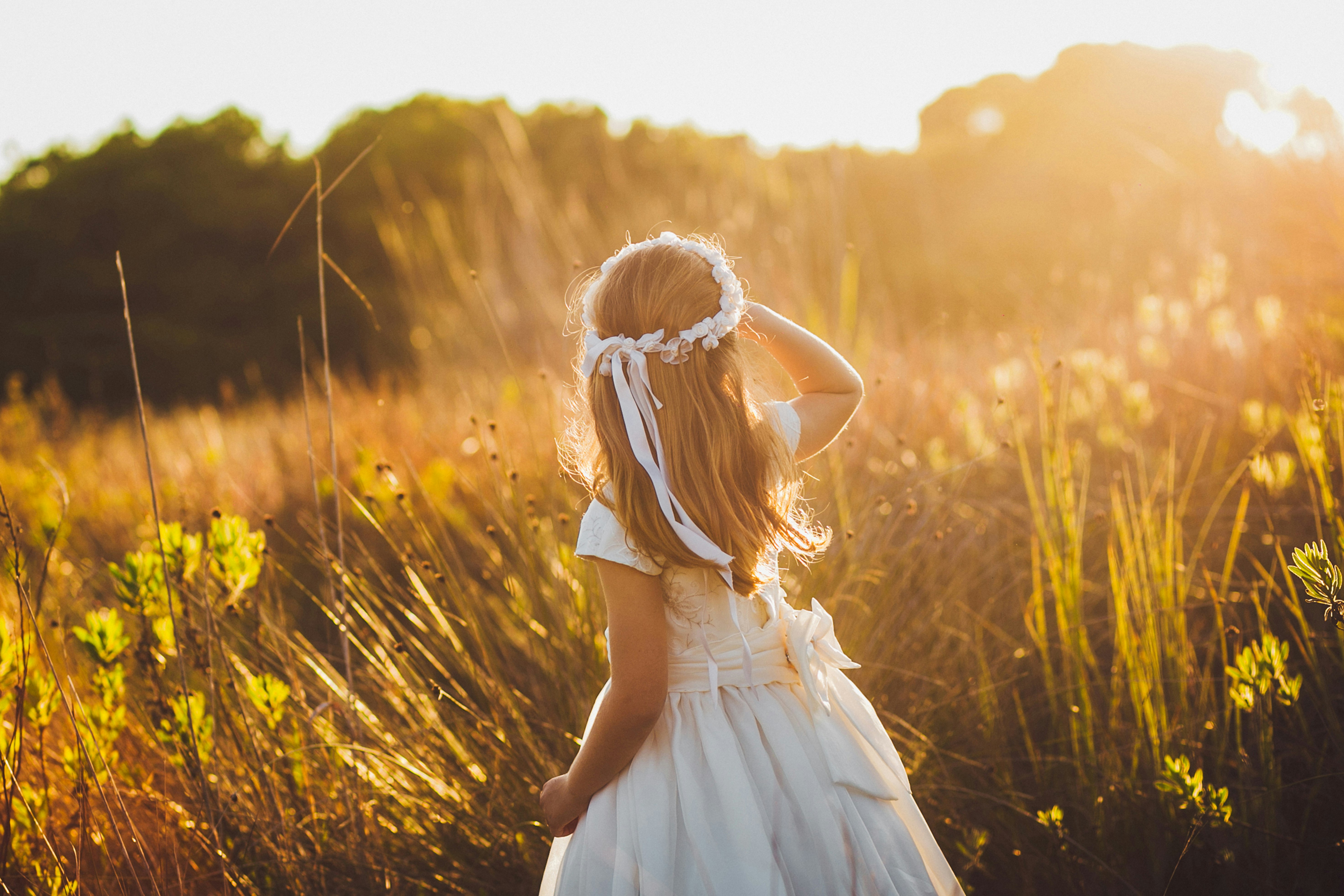 Golden Gates Kindersparplan Ein kleines Mädchen, im weißen Kleid, steht in einer Blumenwiese. Es sieht in die Sonne, welche die Wiese in ein strahlendes Gold taucht. Legen auch Sie Geld für Ihre Kinder richtig an und sparen Sie in Anlagemetalle.