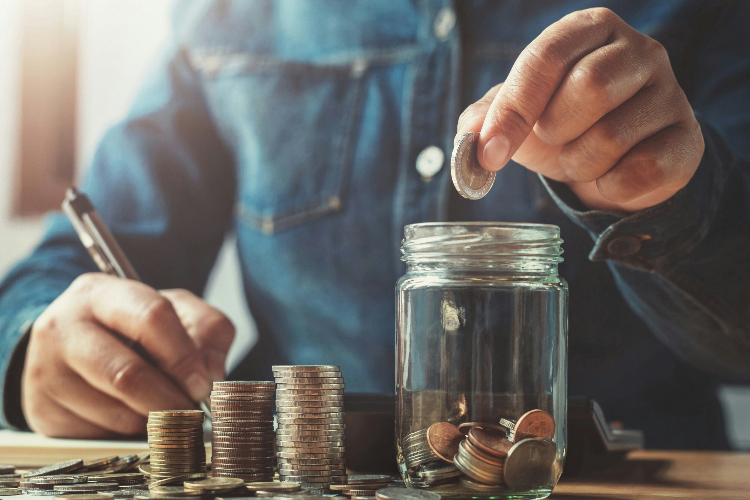 Welche Sparformen sind 2022 noch sinnvoll? In einem hellen Raum sieht man, an einem Tisch eine Person sitzen. Diese zählt Münzen und schreibt die bestände auf. Sparen Sie Ihr Geld mit der Chance auf Wertsteigerung, mit den Edelmetallsparplänen von Golden Gates.