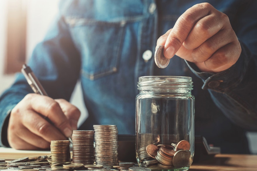 In einem hellen Raum sieht man, an einem Tisch eine Person sitzen. Diese zählt Münzen und schreibt die bestände auf. Sparen Sie Ihr Geld mit der Chance auf Wertsteigerung, mit den Edelmetallsparplänen von Golden Gates.