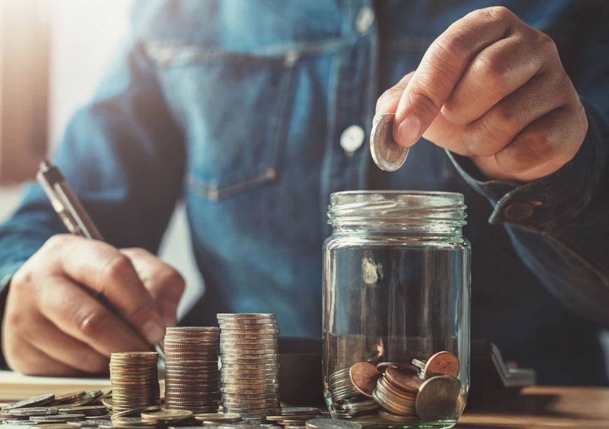 Welche Sparformen sind 2022 noch sinnvoll? In einem hellen Raum sieht man, an einem Tisch eine Person sitzen. Diese zählt Münzen und schreibt die bestände auf. Sparen Sie Ihr Geld mit der Chance auf Wertsteigerung, mit den Edelmetallsparplänen von Golden Gates.