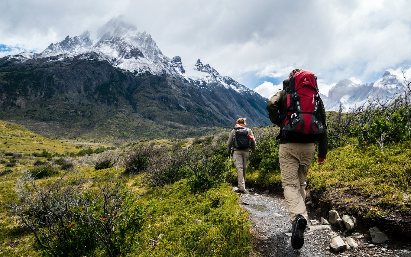 Zwei Personen, die einen Wanderweg entlanglaufen. Alles um sie herum ist grün und im Hintergrund sieht man die Berge mit ihren schneeweißen Bergspritzen. Gehen auch Sie sicher in Ihre Zukunft mit den Edelmetallsparplänen von Golden Gates.