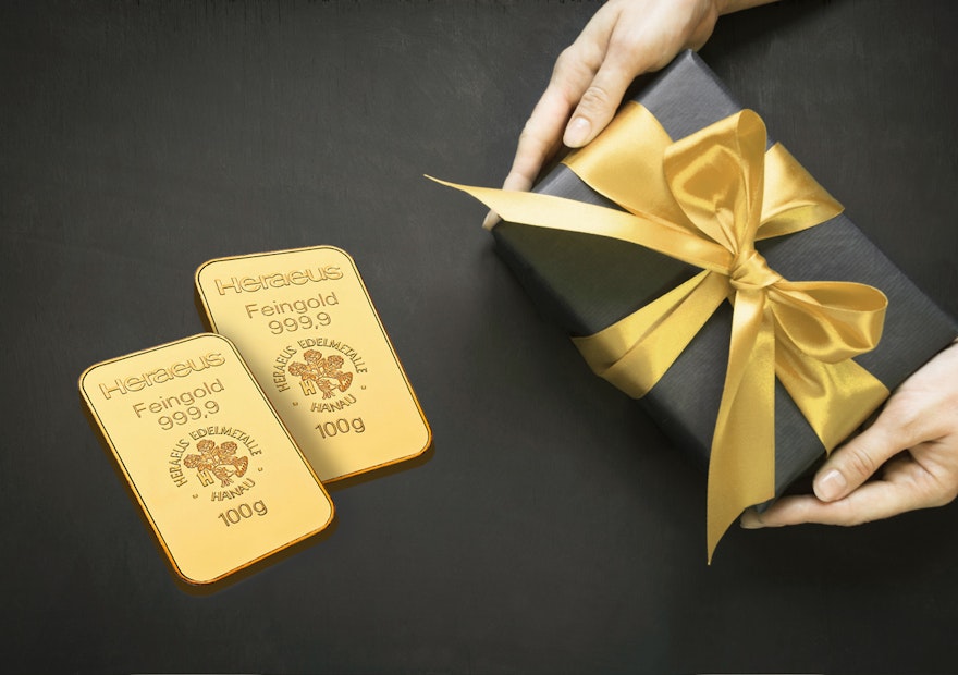 Gold schenken | Golden Gates Auf einem schwarzen Untergrund liegt ein schwarz eingepacktes Geschenk mit goldener Schleife und daneben zwei Goldbarren. Gold für jeden Anlass: Hochzeit, Taufe, Schulanfang, Weihnachten, Geburtstag, mit Golden Gates.