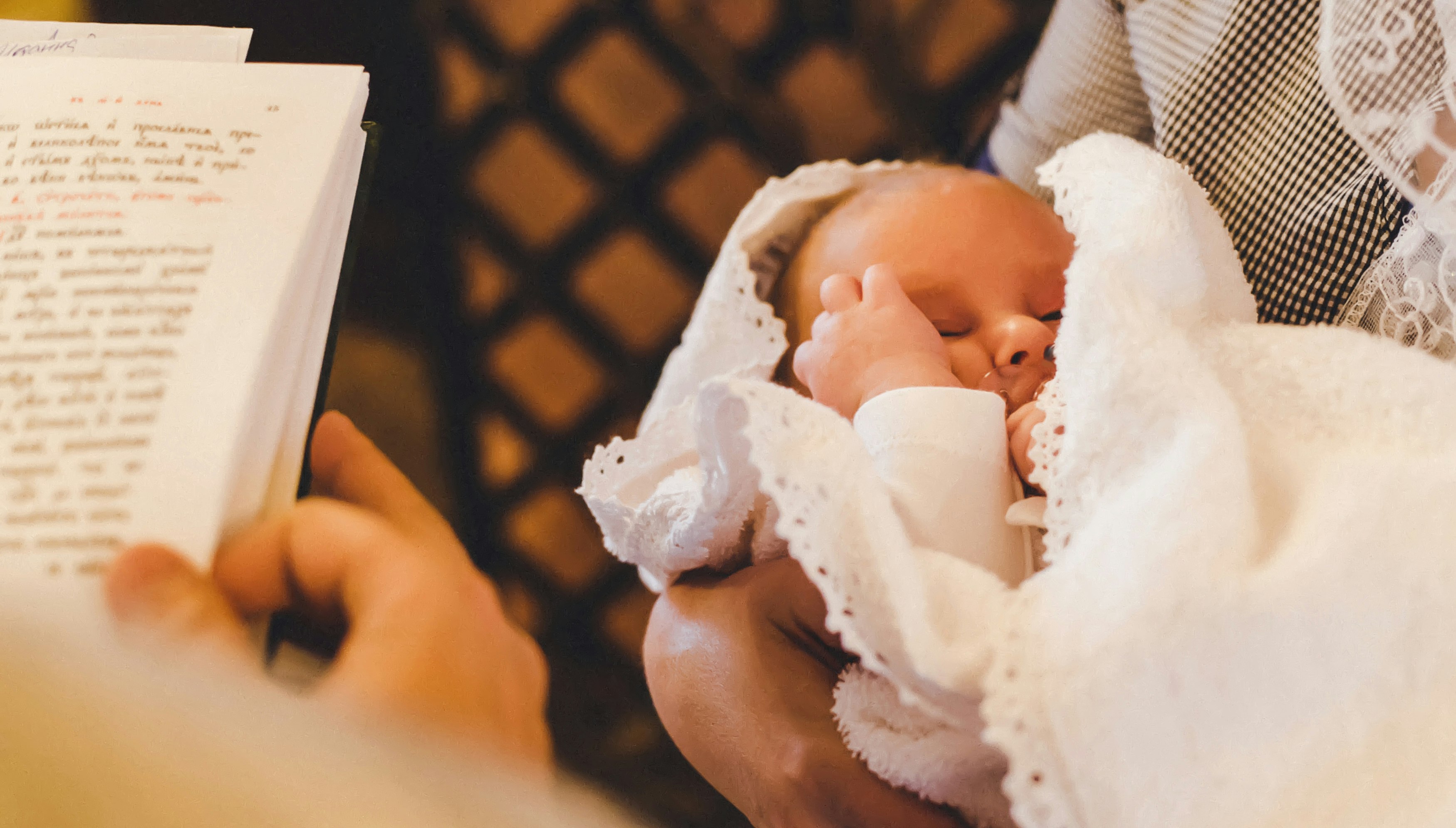 Golden Gates I Geschenke zur Taufe Das Foto wurde über die Schulter des Priesters gemacht, deshalb sieht man einen Teil der Bibel und davor ein Baby in den Armen seiner Mutter. Besondere Momente bedürfen besonderen Geschenken. Mit Golden Gates Gold schenken.