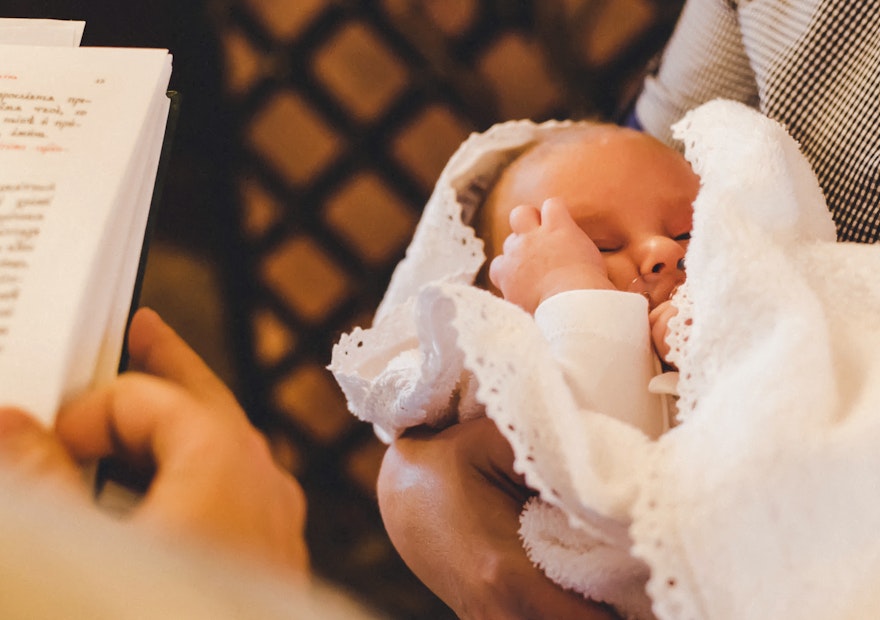 Golden Gates I Geschenke zur Taufe Das Foto wurde über die Schulter des Priesters gemacht, deshalb sieht man einen Teil der Bibel und davor ein Baby in den Armen seiner Mutter. Besondere Momente bedürfen besonderen Geschenken. Mit Golden Gates Gold schenken.