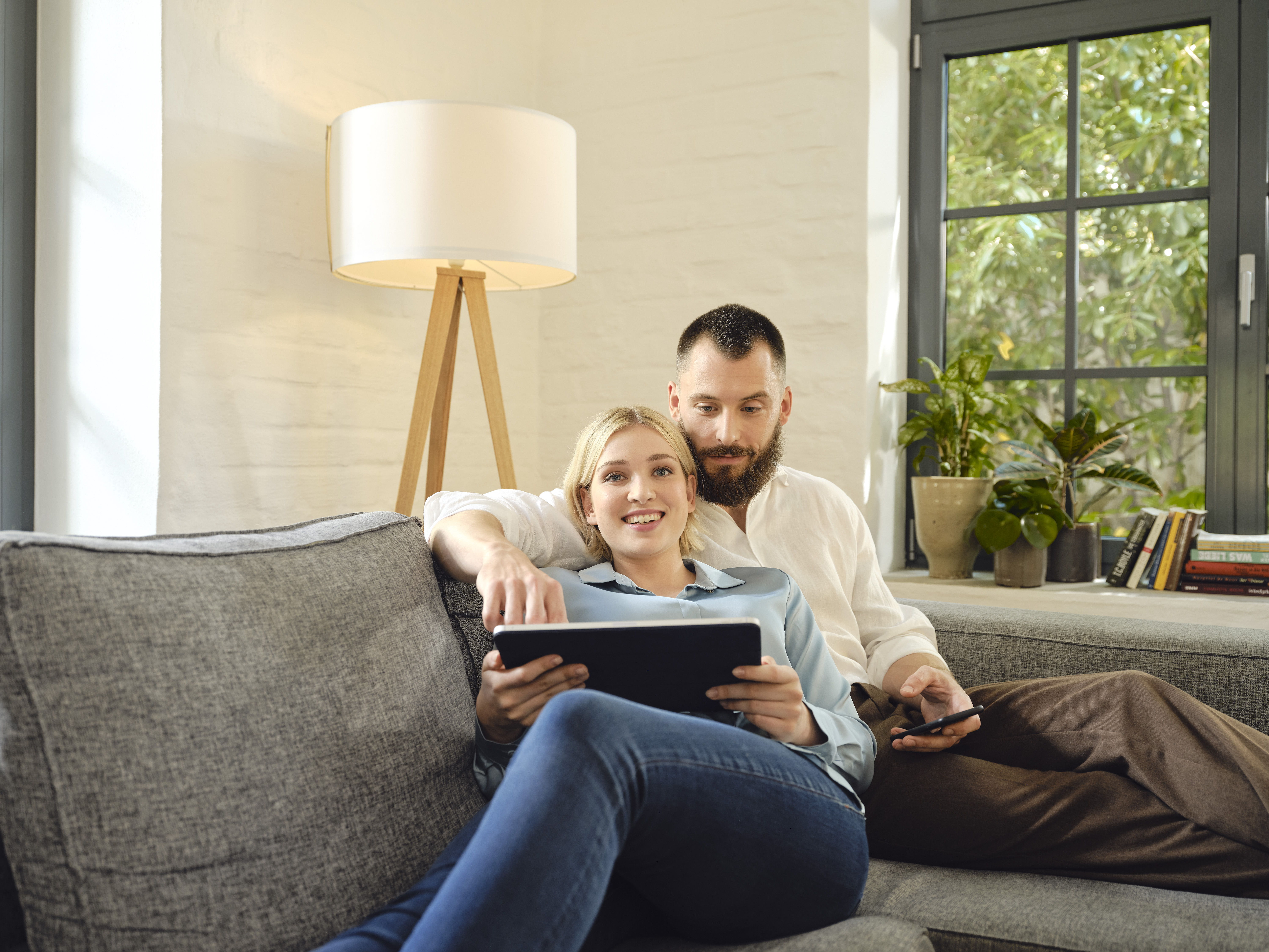 In einem hellen Raum auf einem Sofa, sitzt ein junges Paar mit einem Tablet und sieht freudig in die Kamera. Legen auch Sie Ihr Geld sicher in Anlagemetalle an, mit den Sparplänen von Golden Gates.