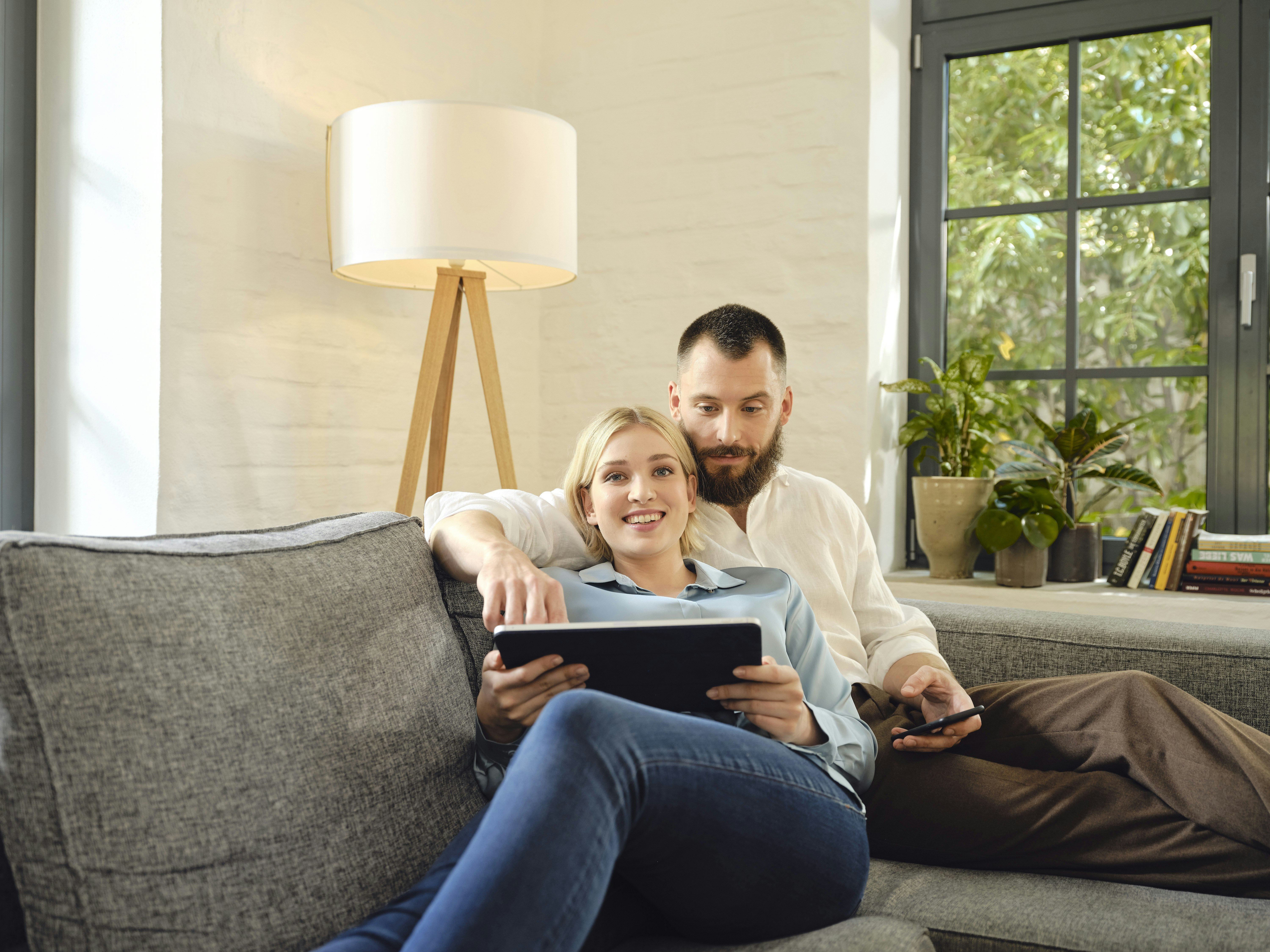 In einem hellen Raum auf einem Sofa, sitzt ein junges Paar mit einem Tablet und sieht freudig in die Kamera. Legen auch Sie Ihr Geld sicher in Anlagemetalle an, mit den Sparplänen von Golden Gates.