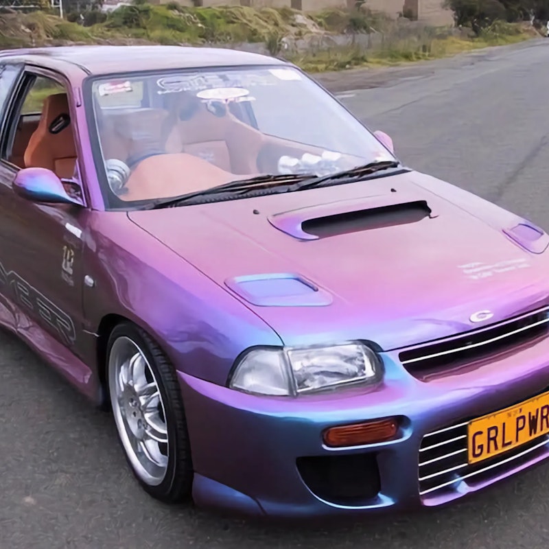 Pink and blue chameleon painted Daihatsu Charade with hood scoop, chrome wheels, and GRLPWR license plate on road