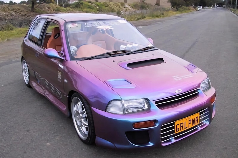 Pink and blue chameleon painted Daihatsu Charade with hood scoop, chrome wheels, and GRLPWR license plate on road