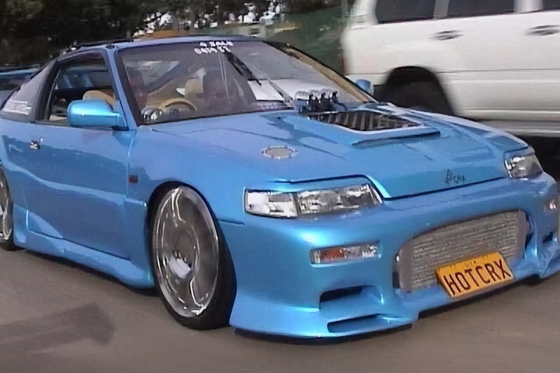 Light blue Honda CRX with exposed blower engine, slammed stance on chrome wheels, and HOTCRX license plate driving on street