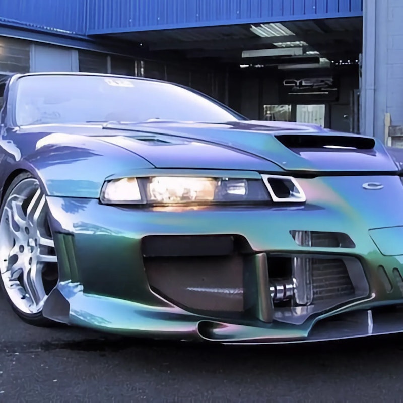 Blue-purple-green chameleon Honda Prelude with wide body kit, aggressive front splitter, and white wheels outside workshop