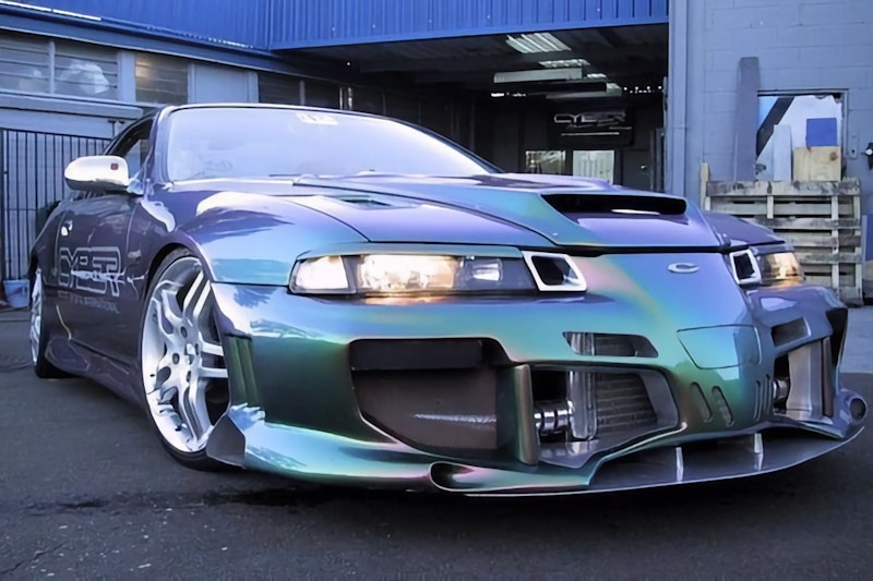 Blue-purple-green chameleon Honda Prelude with wide body kit, aggressive front splitter, and white wheels outside workshop