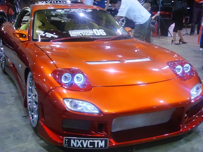 Orange Mazda RX-7 FD with quad headlights, chrome wheels, and NXVCTM plate at show