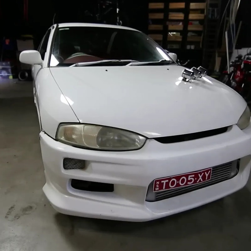 White modified Mitsubishi Lancer with body kit, front lip, intercooler, and red NSW personalised plate "TO·05·XY" photographed in a garage.