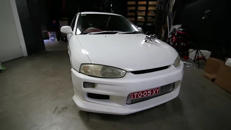 White modified Mitsubishi Lancer with body kit, front lip, intercooler, and red NSW personalised plate "TO·05·XY" photographed in a garage.