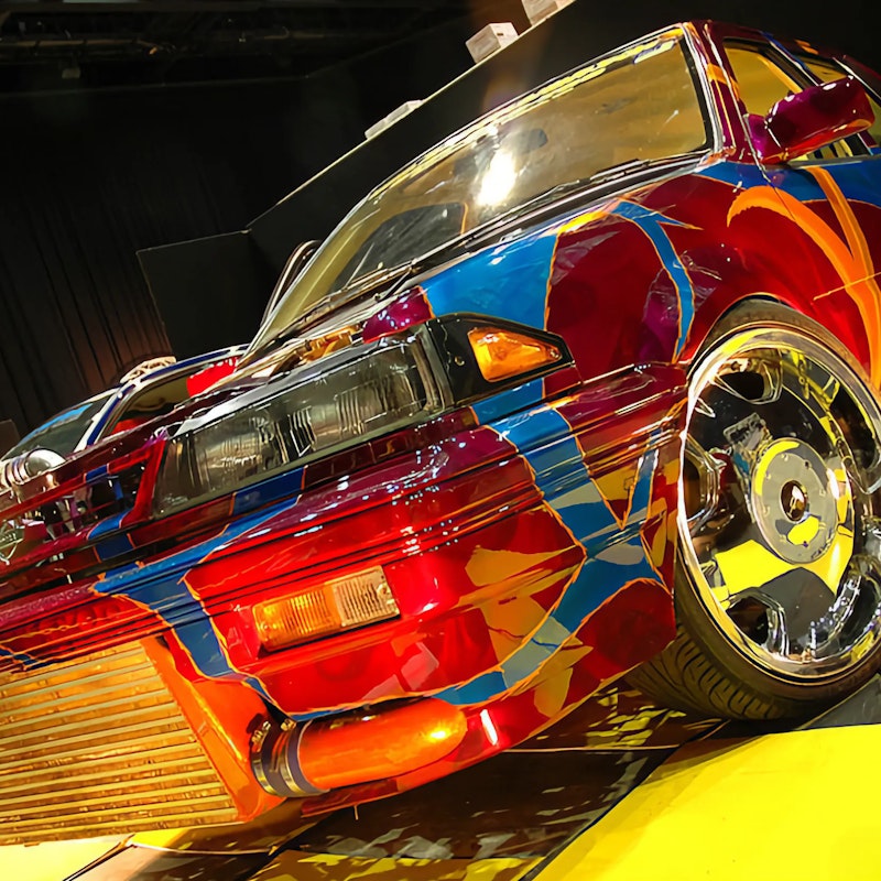 Front of a wildly customised Nissan with multicolour abstract art paintwork, large front-mount intercooler, chrome wheels, and Victorian plate "HVNSNT" on a show display platform.