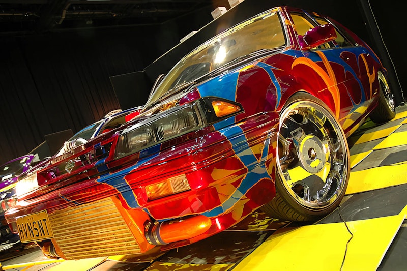 Front of a wildly customised Nissan with multicolour abstract art paintwork, large front-mount intercooler, chrome wheels, and Victorian plate "HVNSNT" on a show display platform.