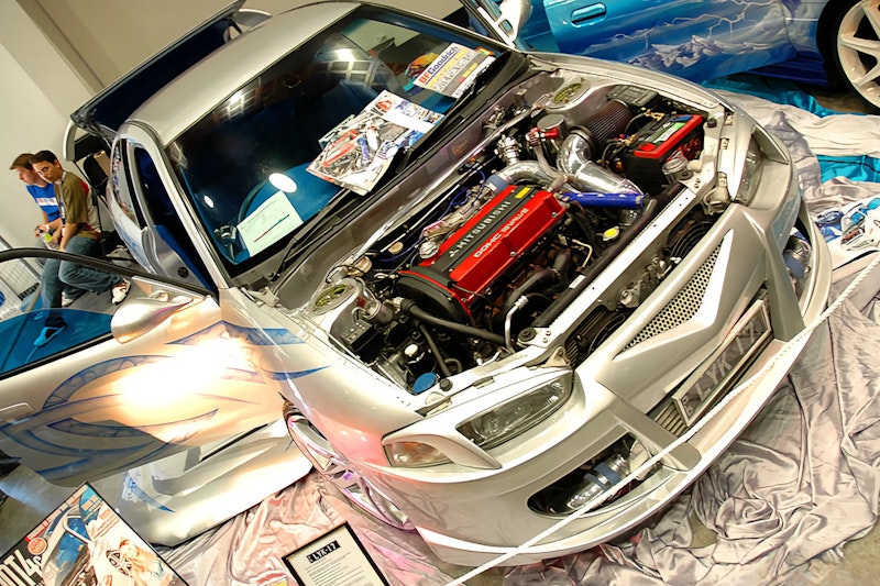 Overhead view of a silver Mitsubishi Lancer Evo at a car show with the bonnet open, revealing a red Mitsubishi DOHC turbo engine with aftermarket intake and piping, custom airbrushed door artwork, and show display materials on the windscreen.