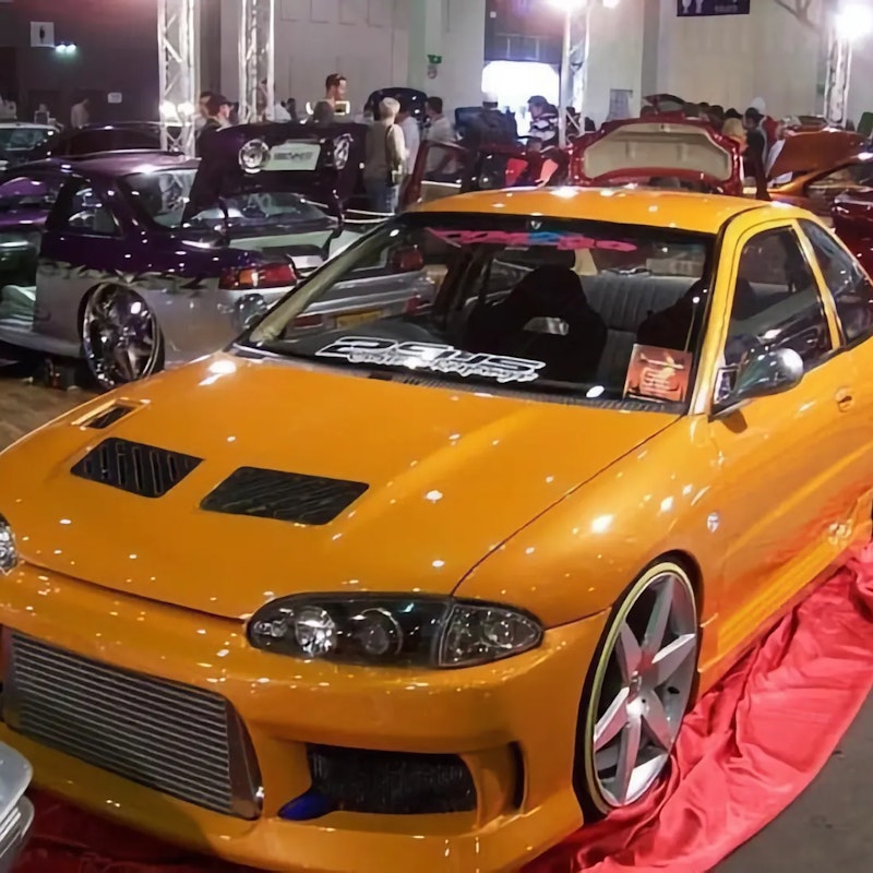 Front three-quarter view of the same orange Lancer on a red carpet at a car show, displaying a large front-mount intercooler, bonnet vents, smoked headlights, and silver wheels.