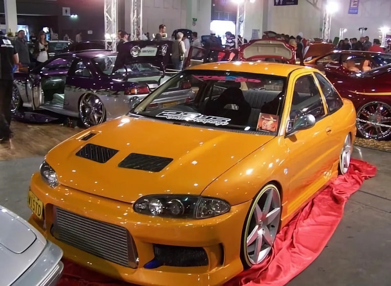 Front three-quarter view of the same orange Lancer on a red carpet at a car show, displaying a large front-mount intercooler, bonnet vents, smoked headlights, and silver wheels.