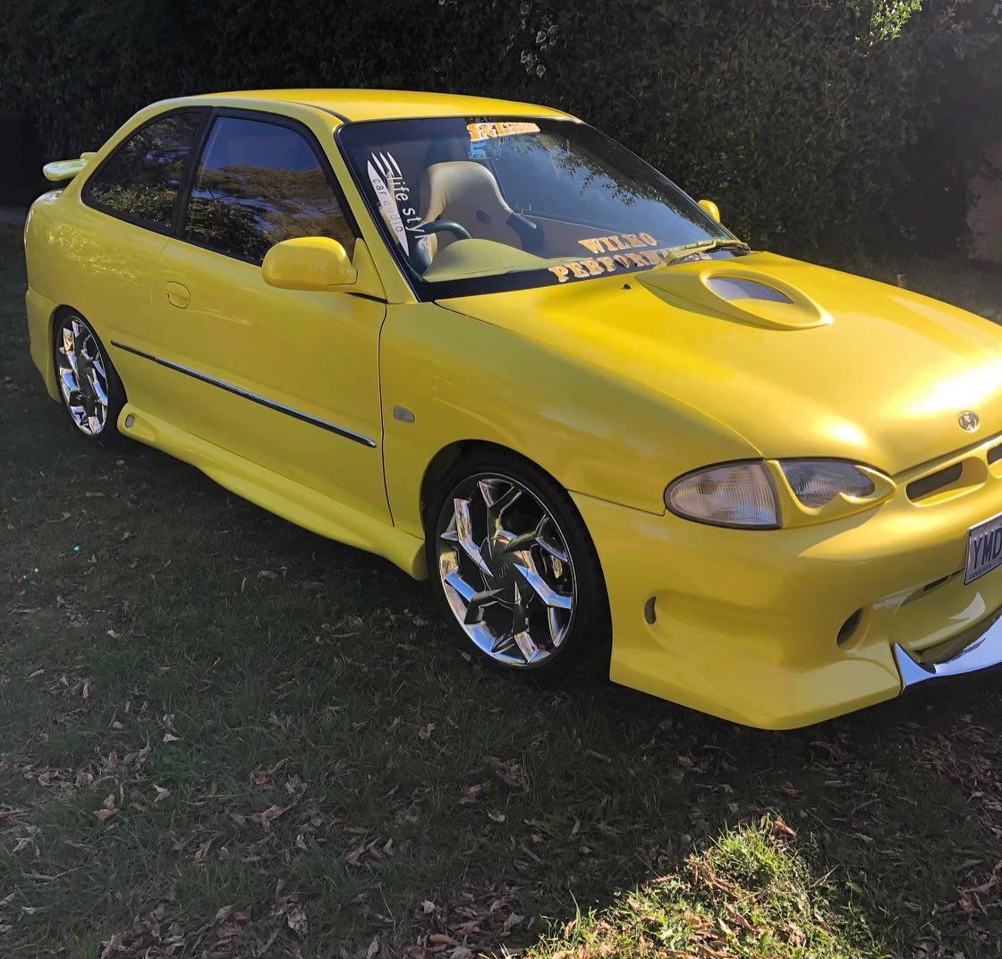 Side-front view of yellow custom Hyundai Excel with chrome wheels and hood scoop