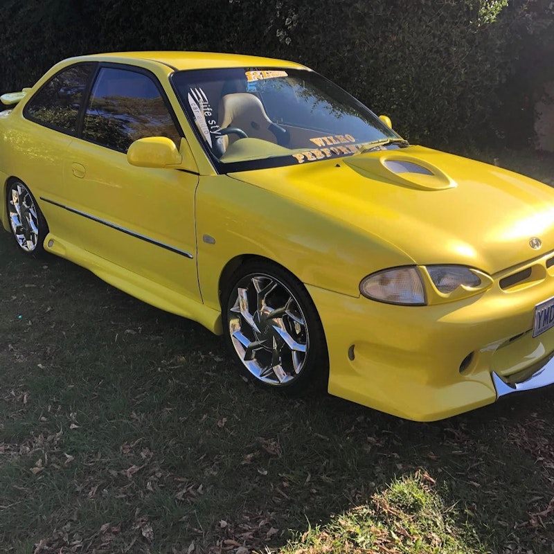 Side-front view of yellow custom Hyundai Excel with chrome wheels and hood scoop