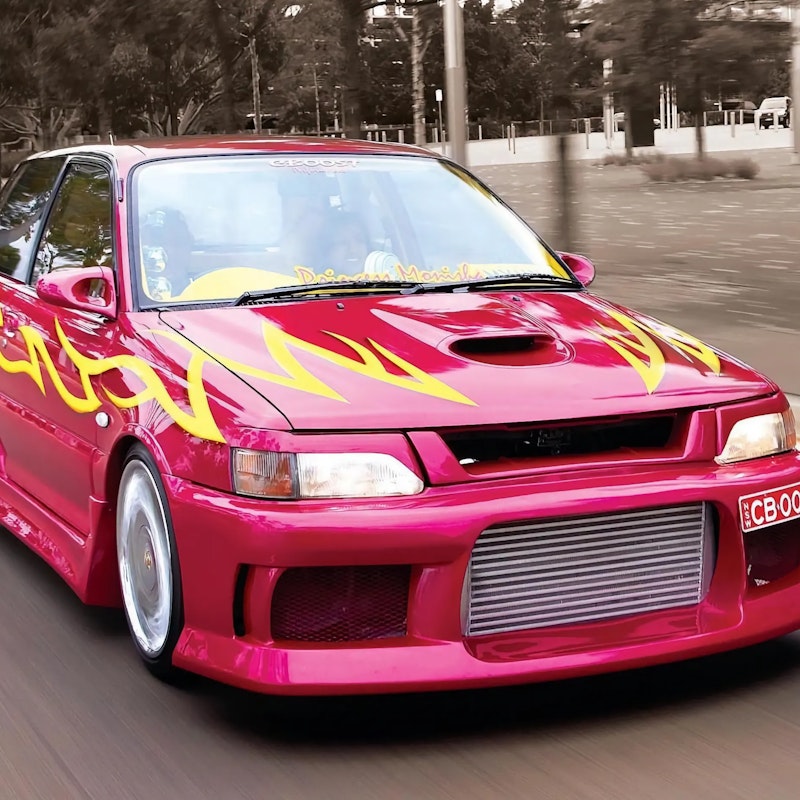 Front three-quarter view of a pink/magenta Toyota Starlet GT Turbo with yellow flame decals and an intercooler-equipped front bumper, driving on a road past a tree-lined median.