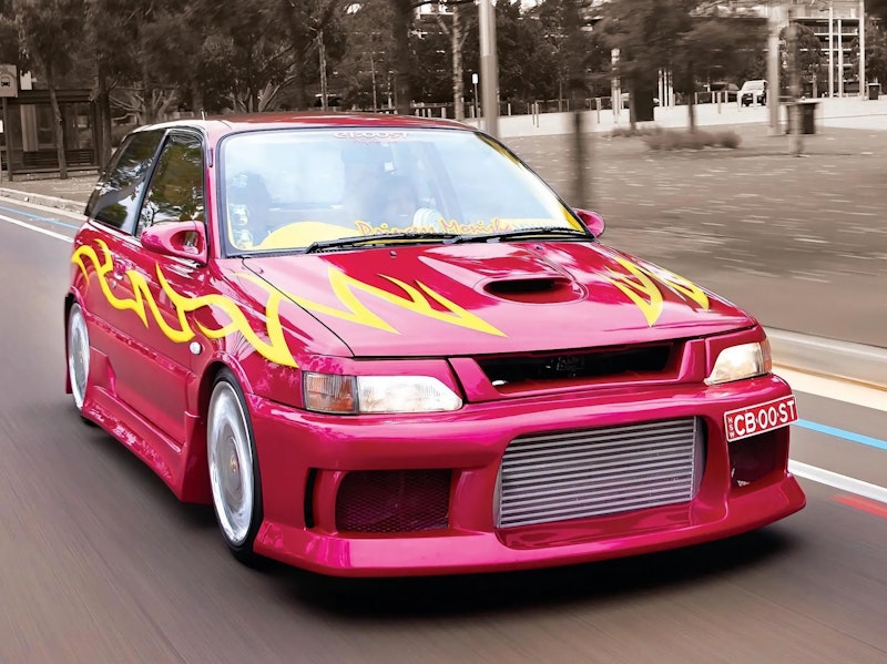 Front three-quarter view of a pink/magenta Toyota Starlet GT Turbo with yellow flame decals and an intercooler-equipped front bumper, driving on a road past a tree-lined median.