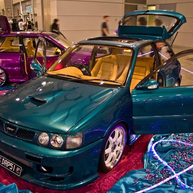 Teal Toyota Starlet GT with flame paint and tan leather interior on display at a car show, doors and hatch open, with a purple Starlet visible behind it.