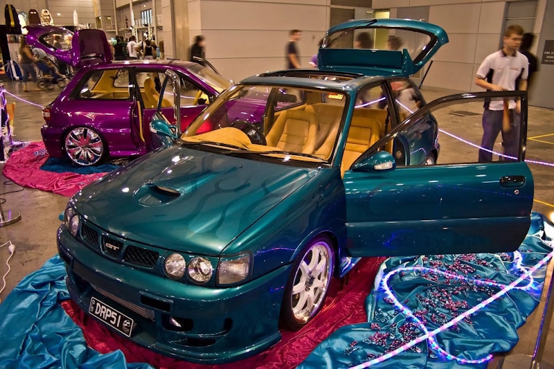 Teal Toyota Starlet GT with flame paint and tan leather interior on display at a car show, doors and hatch open, with a purple Starlet visible behind it.