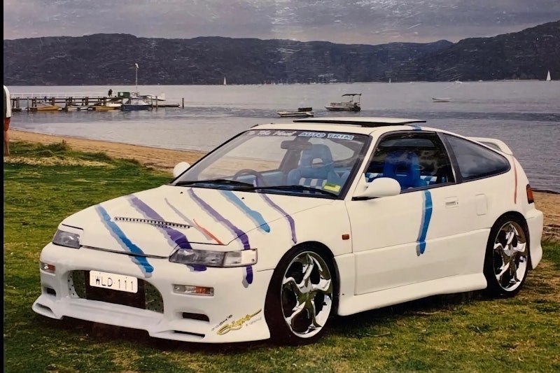 Front three-quarter view of the white Honda CRX with claw-mark graphics, parked on grass beside a lake with boats and hills in the background.