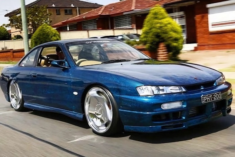 Front three-quarter view of a metallic blue Nissan Silvia S14 with deep-dish chrome wheels, driving on a suburban street past brick houses.