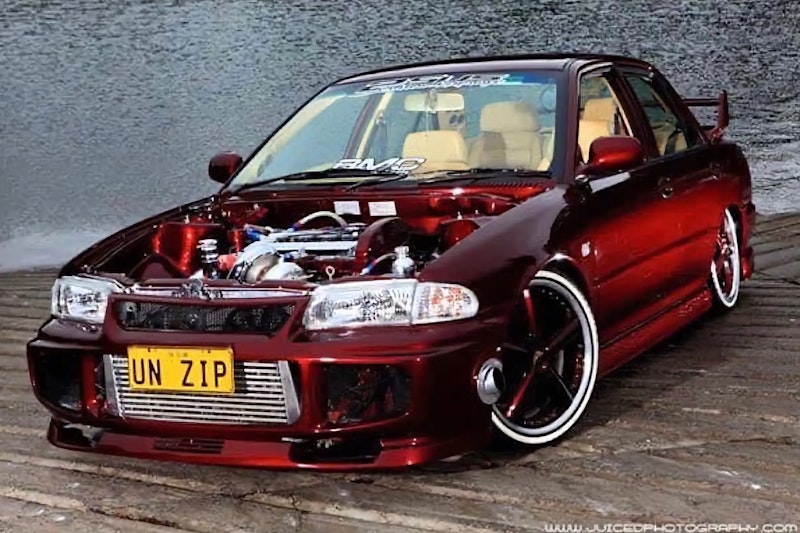 Front three-quarter view of a deep red Mitsubishi Lancer Evolution with its bonnet removed showing a detailed turbocharged engine bay, custom wheels, and "UN ZIP" number plate, parked on a wooden dock by water.