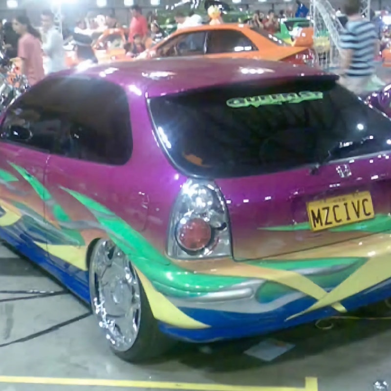 Rear three-quarter view of a purple Honda Civic hatchback with multicolor flame graphics and "MZCIVC" number plate on display at an indoor car show, with other modified cars and a crowd in the background.