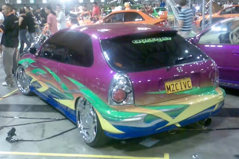 Rear three-quarter view of a purple Honda Civic hatchback with multicolor flame graphics and "MZCIVC" number plate on display at an indoor car show, with other modified cars and a crowd in the background.