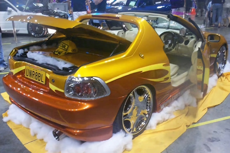 Rear three-quarter view of a custom gold and orange Honda CR-X del Sol with flame graphics, gold-and-chrome wheels, and "UNR8BL" number plate, displayed on a car show stand with the boot open and cotton-cloud styling.