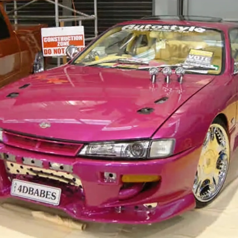 Front three-quarter view of a pink/magenta Nissan Silvia S14 with a custom bumper, chrome wheels, and "4DBABES" number plate, on display indoors next to an orange custom ute.