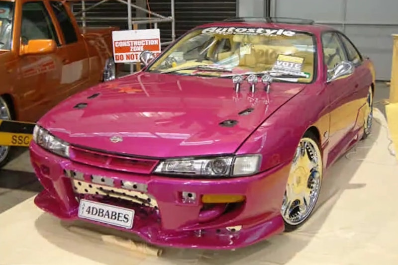 Front three-quarter view of a pink/magenta Nissan Silvia S14 with a custom bumper, chrome wheels, and "4DBABES" number plate, on display indoors next to an orange custom ute.