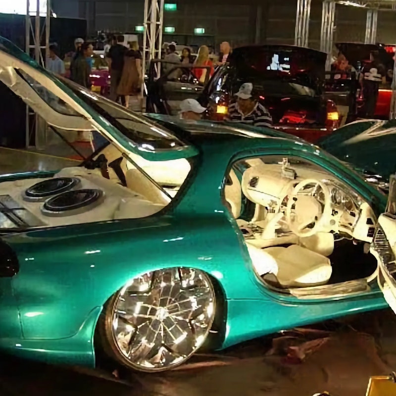 Green Mazda RX-7 with boot, doors, and bonnet open displaying a white custom interior and boot-mounted speakers, on show at an indoor car event.