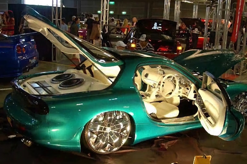 Green Mazda RX-7 with boot, doors, and bonnet open displaying a white custom interior and boot-mounted speakers, on show at an indoor car event.