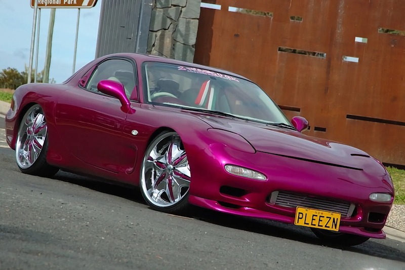 Front three-quarter view of a magenta Mazda RX-7 with "PLEEZN" number plate, parked near a wooden fence.