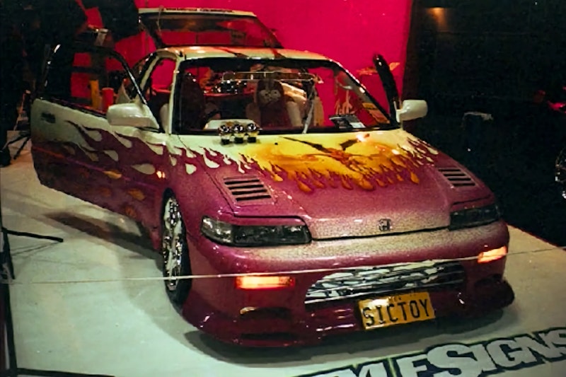 Front three-quarter view of a Honda CRX with white/maroon flame paint and "SICTOY" number plate, displayed indoors at a car show with doors open.