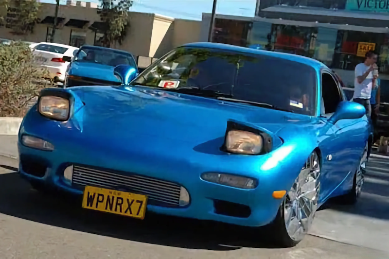 Front three-quarter view of a blue Mazda RX-7 with pop-up headlights raised, driving through a car park near shops.