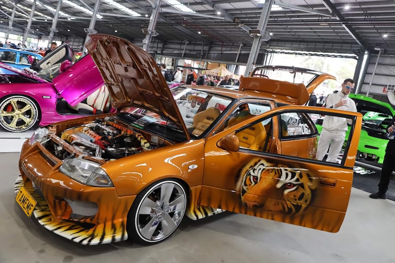 Side view of an orange tiger-livery Suzuki Swift with bonnet and door open, showing the engine bay and a tiger face airbrushed on the door, parked among other show cars.