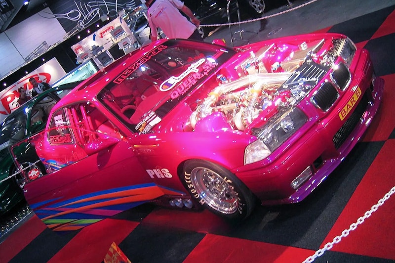 Front three-quarter view of a pink BMW E36 with bonnet open showing a chrome-piped turbo engine, drag wheels, rainbow side graphics, and roll cage, on a checkered show floor.
