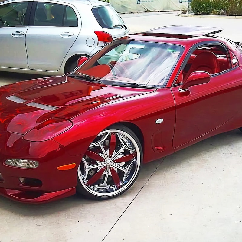 Front three-quarter view of a red Mazda RX-7 with flame graphics on the bonnet and chrome wheels with red inserts, parked outdoors next to a silver hatchback.